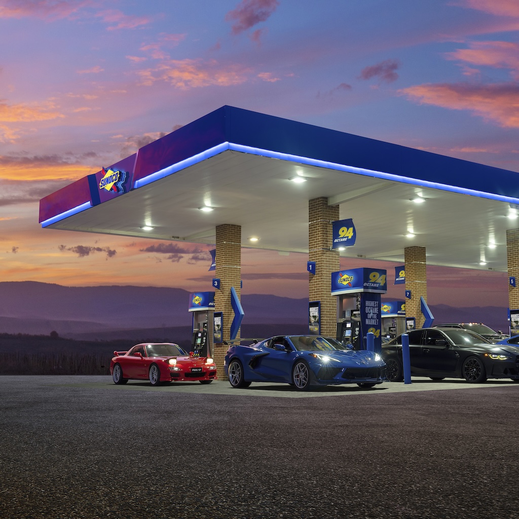 Three sports cars parked under a lit Sunoco canopy during a colorful sunset, evoking the nostalgia, routine, and excitement that fuel road trip rituals and station loyalty