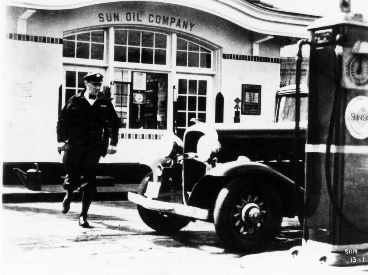 Vintage Sunoco gas station with attendant and classic car, circa early 20th century.